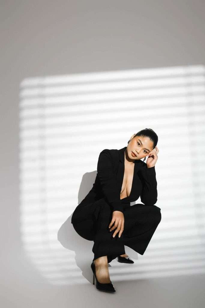 Woman in a black suit squats on the floor, looking at the camera, with striped light and shadow pattern on the wall behind her.