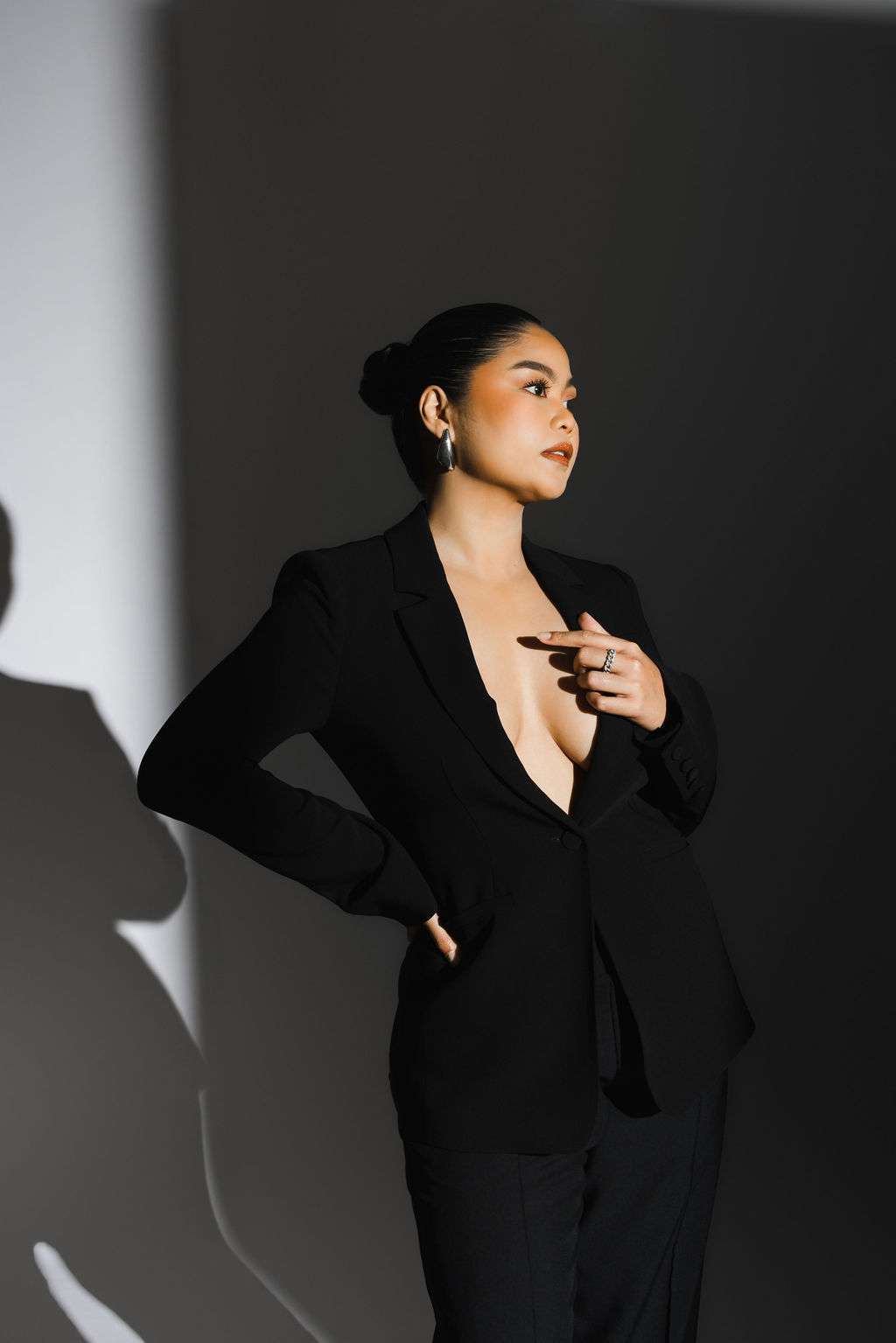 Person in a black suit with deep neckline stands against a shadowed wall, looking to the side with one hand on hip and the other touching their chest.