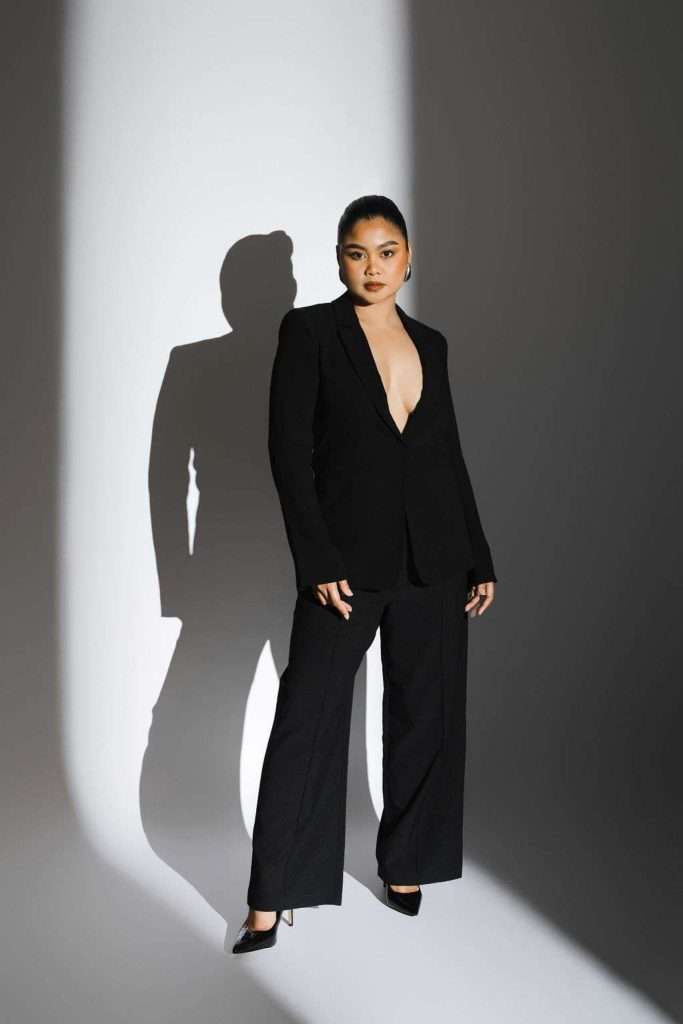 Person wearing a black suit with no shirt stands confidently in a spotlight, casting a shadow on a white and gray background.