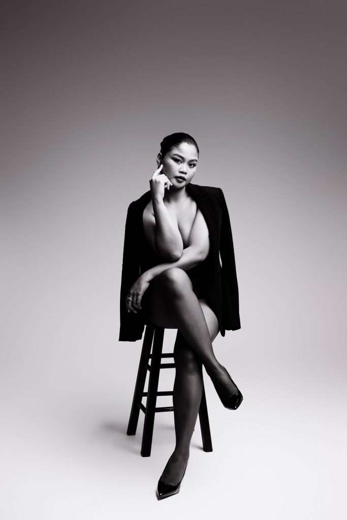 A woman sits on a stool wearing a blazer and heels, crossing her legs and resting her chin on her hand, against a plain gradient background.