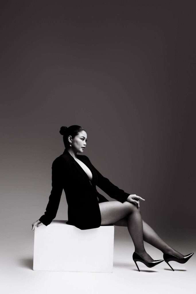 Woman in a black blazer and high heels sits on a white block against a plain background, looking to the side.