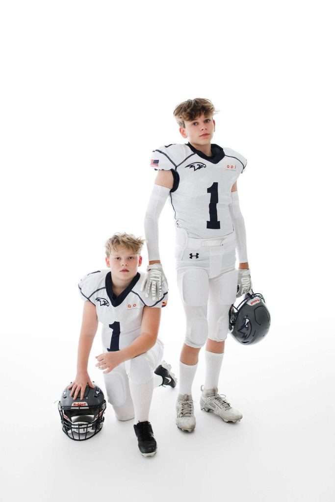 Two boys in white football uniforms, each wearing the number 1, pose in a studio; one is kneeling with a helmet, while the other stands, both looking at the camera against a white background.