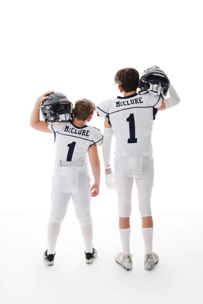 Two youth football players in white uniforms with "McClure" and the number 1 on their backs stand side by side, holding helmets, facing away from the camera against a white background.