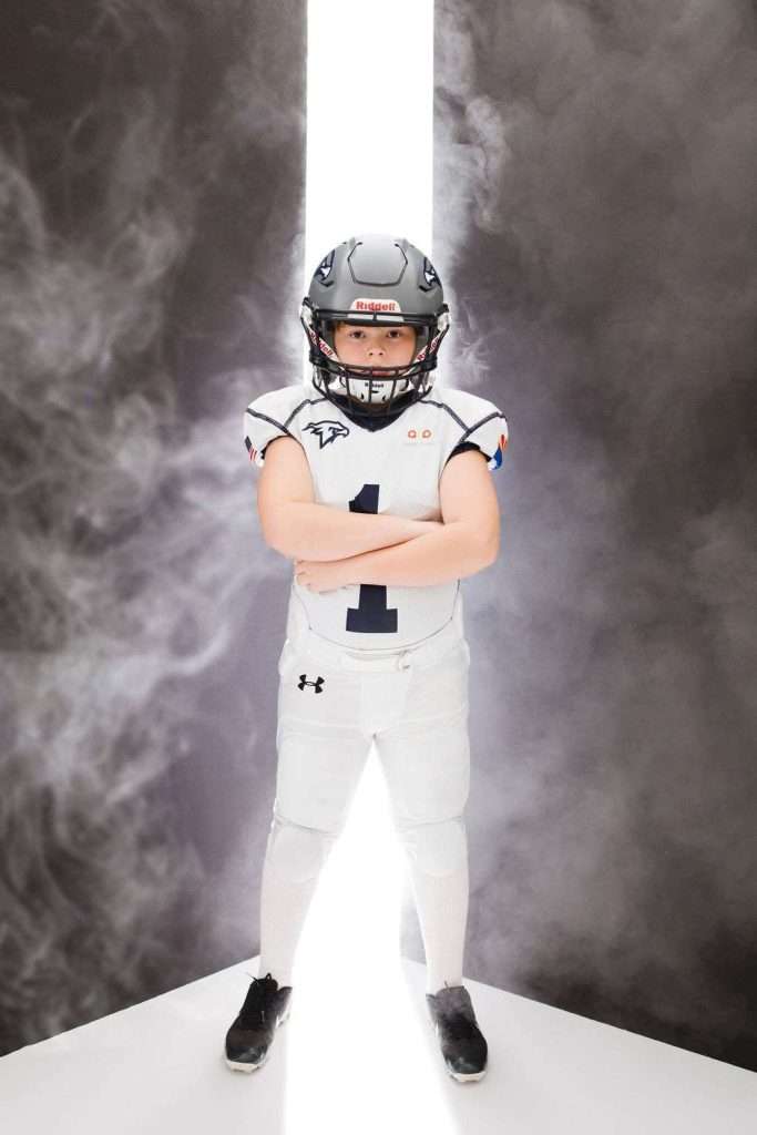 Young football player in full uniform and helmet stands with arms crossed against a dramatic, smoky background with a bright vertical light.