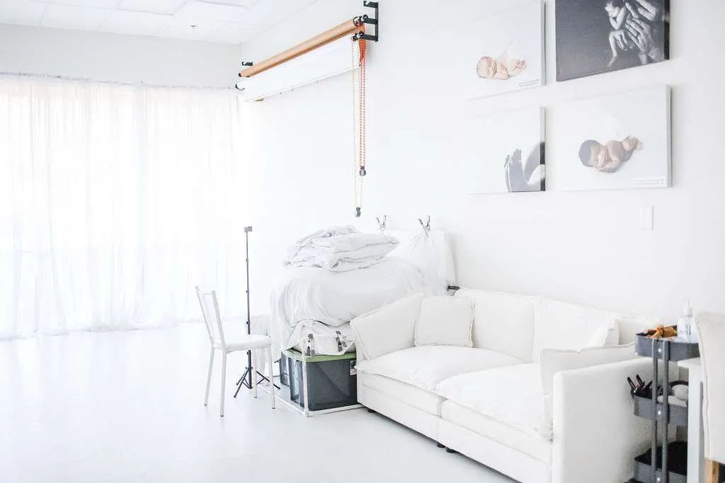 Bright, minimalist photo studio with white walls, a white sofa, stacked bedding, photography equipment, and newborn portraits on the wall.