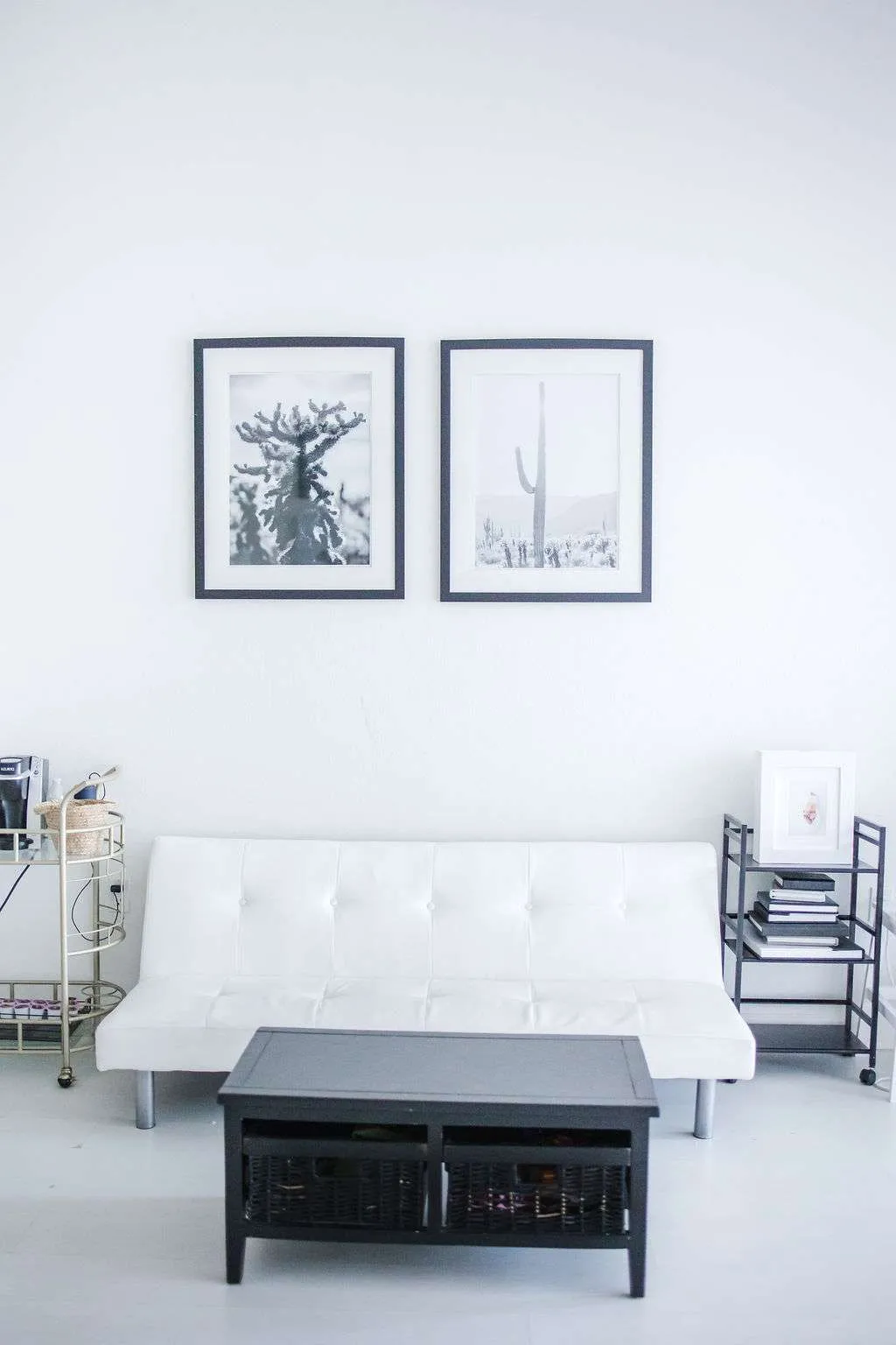 Minimalist living room with a white futon, black coffee table, side carts with decor, and two framed cactus photos on a white wall.