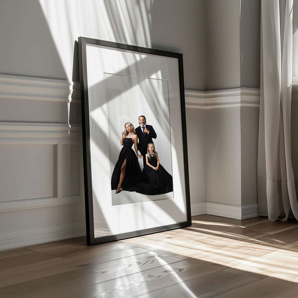Framed portrait of three adults in formal black attire resting against a sunlit wall in a minimalist room with wooden floors and white paneling.
