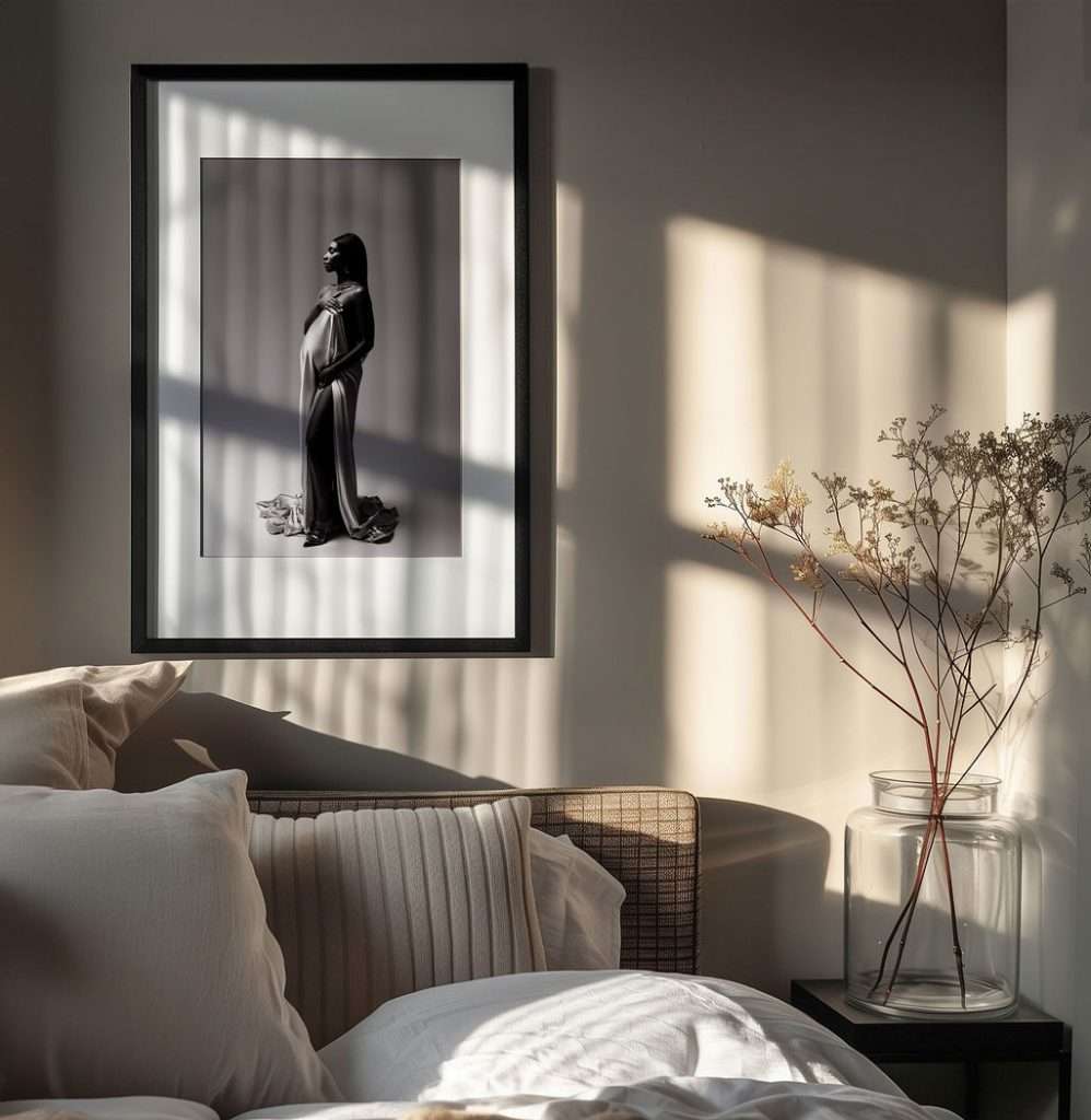 A framed black and white photo of a woman in a gown hangs above a couch with white pillows; sunlight casts shadows on the wall and a glass vase with dried flowers sits nearby.