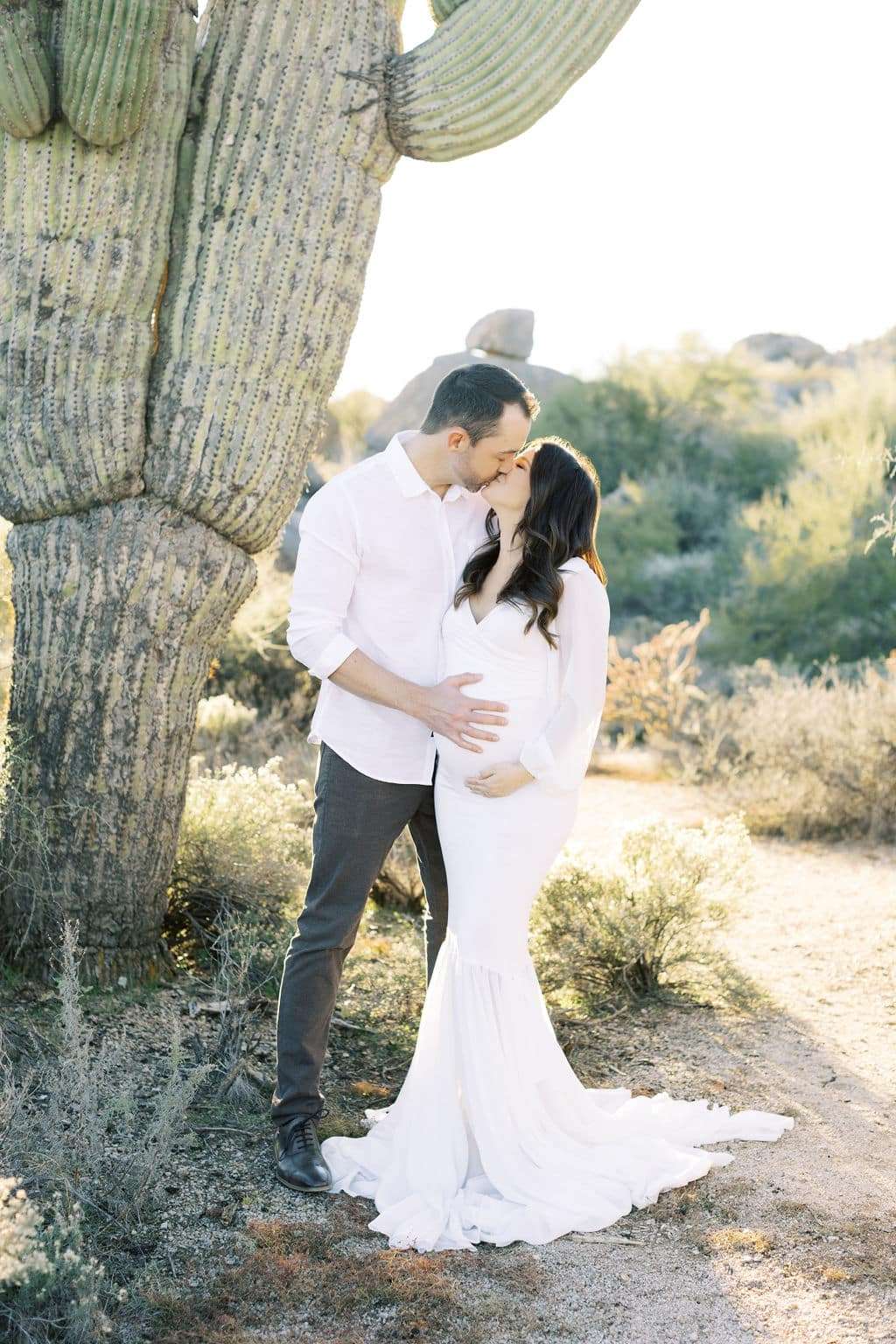 A couple stands by a large cactus in the desert. The man kisses the woman’s forehead as she cradles her pregnant belly. Both are dressed in light-colored clothing.